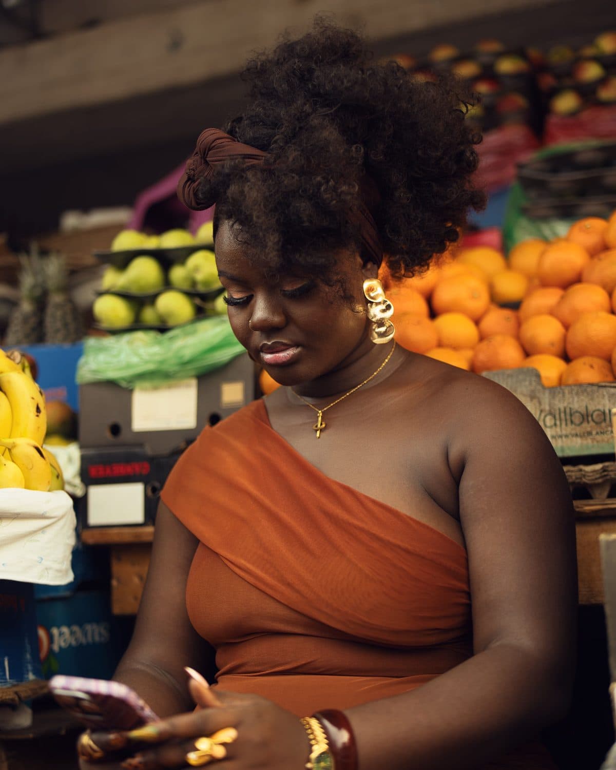 Au marché, une cliente regarde son téléphone, devant des étals de fruits.